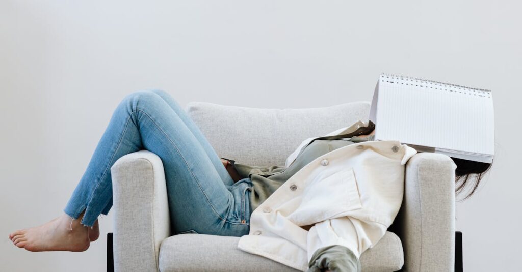 Tired woman in casual attire falls asleep on sofa with a notebook on face and pen in hand.